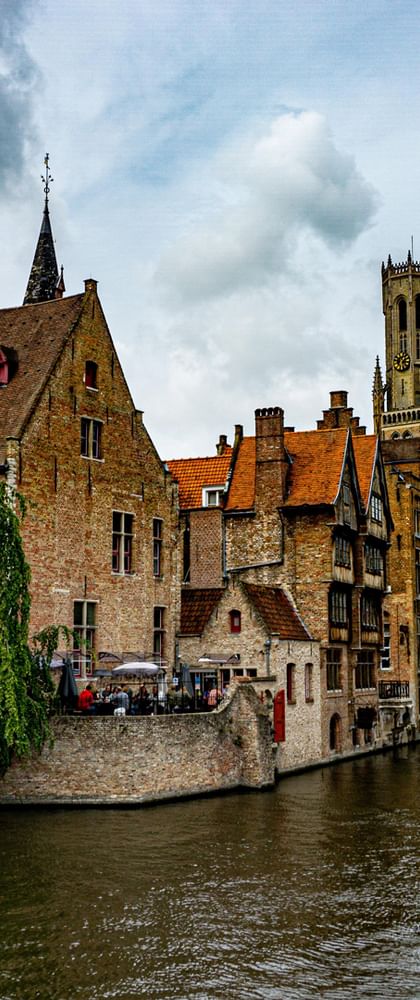 Mittelalterliche Gebäude mit Treppengiebeln und der Belfried-Turm entlang eines Kanals in Brügge. Touristenboote fahren unter bewölktem Himmel.