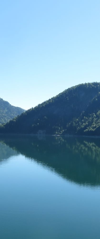 Sylvensteinsee mountain lake with perfect mirror reflection of forested hills under clear blue sky in Bavaria, Germany.