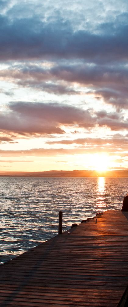 Zwei Personen sitzen auf einem Holzsteg, der ins ruhige Wasser führt bei Sonnenuntergang. Dramatische Wolken in Orange und Blau füllen den Himmel in Valencia.