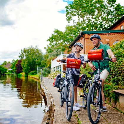Two cyclists with helmets and red panniers on bikes beside a canal in Hinte, East Frisia. Traditional brick buildings and green trees visible.
