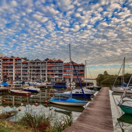 Hafen in Rastede mit Segelbooten am Holzsteg. Moderne Wohngebäude säumen das Ufer unter dramatischem Wolkenhimmel bei Sonnenuntergang.