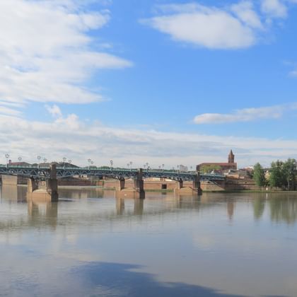 Steinbrücke über den Canal du Midi in Toulouse mit historischen Gebäuden und grüner Kuppel am Flussufer unter blauem Himmel.