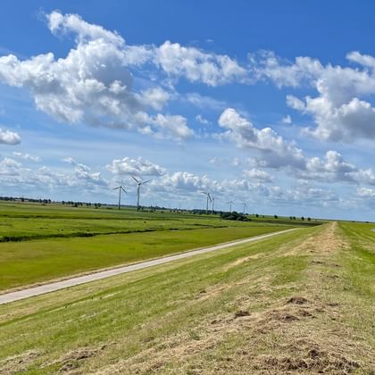 Two cyclists on a paved path along a grass-covered dike in East Frisia. Wind turbines visible in the distance under a blue sky with clouds.
