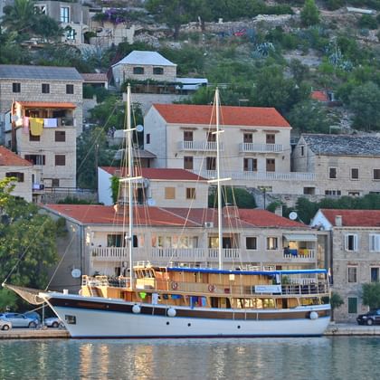 Traditionelles Holzschiff San Snova mit zwei Masten im Hafen in Kroatien, mit Steinhäusern und grünem Hang im Hintergrund.