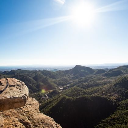 Zwei Radfahrer in roten Jacken auf Mountainbikes am felsigen Klippenrand mit Blick auf bewaldete Berge und Täler in der Region Valencia, Spanien.