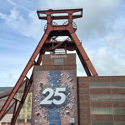 Roter Förderturm der Zeche Zollverein mit Backsteingebäude und '25'-Banner. Die ikonische Industriestruktur erhebt sich vor blauem Himmel.