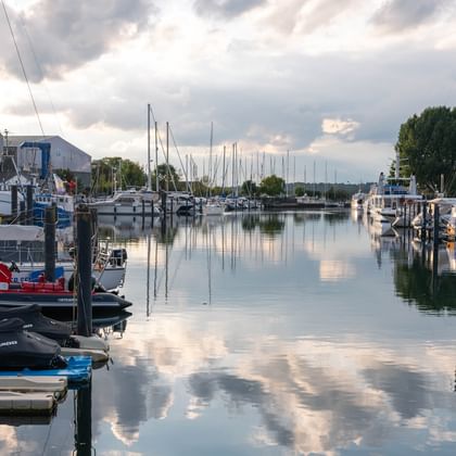 Ruhiger Yachthafen in Travemünde mit verschiedenen Booten an Holzstegen. Das ruhige Wasser spiegelt bewölkten Himmel und Masten perfekt wider.
