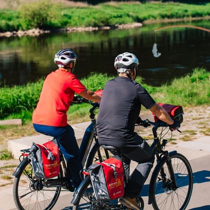 Zwei Radfahrer mit Helmen und Tourenrädern mit roten Packtaschen sitzen auf Kopfsteinpflaster an der Weser. Im Hintergrund angelt eine Person.