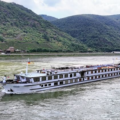 White river cruise ship MS Veloria with German flag sailing on the Rhine River, with green vineyard-covered hills in the background.