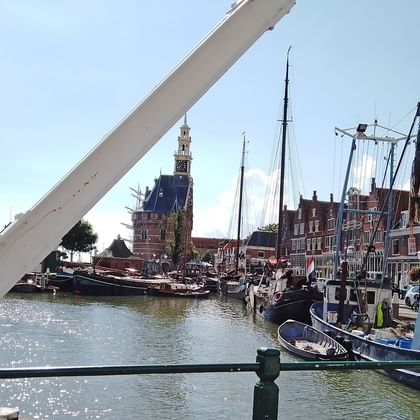 Niederländischer Hafen mit historischen Segelschiffen am Kanal. Traditionelle Gebäude und Kirchturm im Hintergrund unter blauem Himmel.