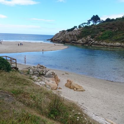 Küstenszene in der Nordbretagne mit einem Sandstrand, wo ein Fluss auf das Meer trifft, mit bewachsenen Felsklippen und spazierenden Menschen.
