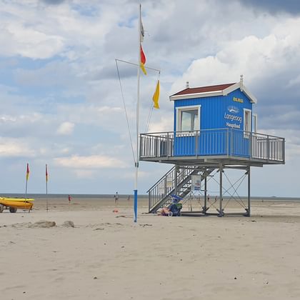 Blaue erhöhte Rettungsstation auf Stelzen am Strand von Langeoog mit deutschen Flaggen, Treppe und gelbem Boot in der Nähe auf sandigem Ufer.