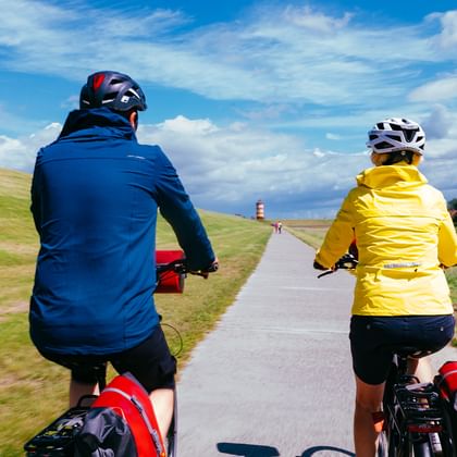 Zwei Radfahrer mit Packtaschen fahren auf gepflastertem Deichweg in Ostfriesland. Einer trägt blaue, der andere gelbe Jacke. Rot-weißer Leuchtturm Pilsum voraus sichtbar.