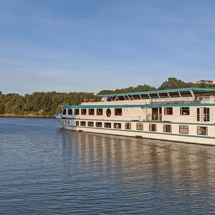 Flusskreuzfahrtschiff MS Switzerland mit weißem Rumpf und grünen Akzenten auf ruhigem Fluss, umgeben von Bäumen unter blauem Himmel.