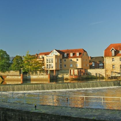 Flussblick auf Rheine mit Wehr, historischen Gebäuden einschließlich Kirchturm, bunten Häusern entlang der Uferpromenade unter blauem Himmel.