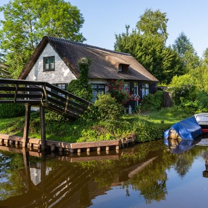 Traditionelles Reetdachhaus neben einem Kanal in Giethoorn mit Holzbrücke und Booten am Ufer, umgeben von üppigem Grün.