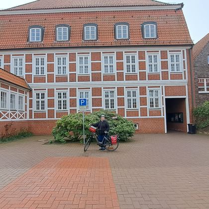 Frau namens Olga mit Fahrrad vor historischem rotem Backsteinrathaus in Bad Bederkesa mit weißen Fenstern und Ziegeldach.
