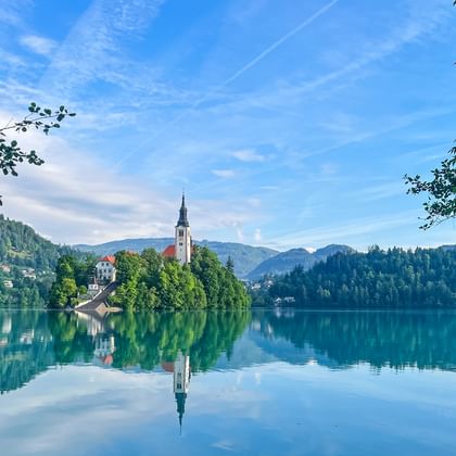 Bleder See in Slowenien mit kleiner Insel mit Kirche und weißem Glockenturm, umgeben von bewaldeten Hügeln und Bergen unter blauem Himmel.
