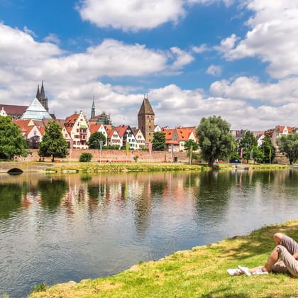 Zwei Radfahrer ruhen im Gras an der Donau in Ulm, mit dem gotischen Ulmer Münster und historischen Gebäuden im Wasser gespiegelt.