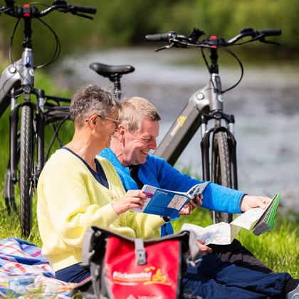 Zwei Radfahrer sitzen im Gras und lesen eine Karte während einer Picknicpause. E-Bikes stehen in der Nähe, Fluss und grüne Landschaft im Hintergrund.