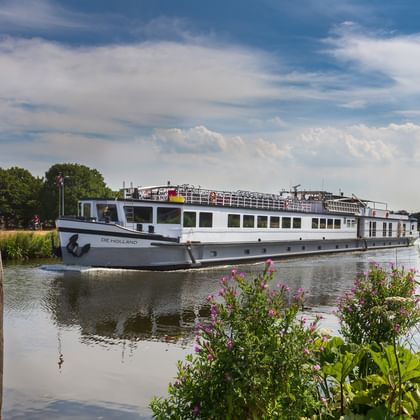 Weißes Flusskreuzfahrtschiff De Holland fährt auf ruhigem Wasser mit hölzernen Dalben im Vordergrund und blühenden Pflanzen am Ufer.