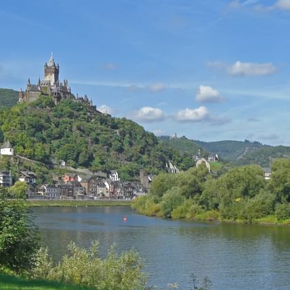 Burg Cochem thront auf einem bewaldeten Hügel über der Mosel, mit der Stadt Cochem am Flussufer darunter.