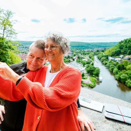 Glückliches Paar macht Selfie mit rotem Handy an malerischem Aussichtspunkt über Hann. Münden mit Weser und grünen Hügeln im Hintergrund.