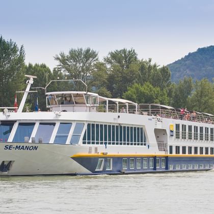 Weißes Flusskreuzfahrtschiff SE-Manon fährt auf der Donau mit Passagieren an Deck, bewaldete Hügel und grünes Flussufer im Hintergrund.