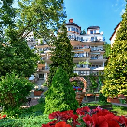 Mehrstöckiges Hotelgebäude mit Balkonen, umgeben von üppigen grünen Gärten mit roten Blumen, alten Bäumen und gepflegten Anlagen.