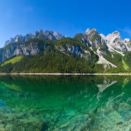 Kristallklarer Gosausee im Salzkammergut mit smaragdgrünem Wasser, das dramatische Alpengipfel unter blauem Himmel spiegelt.