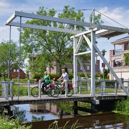 Zwei Radfahrer mit Rädern stehen auf einer weißen Metall-Zugbrücke über einem Kanal. Traditionelle holländische Häuser und grüne Bäume im Hintergrund.