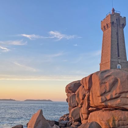 Steinerner Leuchtturm auf rosa Granitfelsen an der Küste bei Sonnenuntergang. Das Meer und entfernte Inseln sind unter blauem Himmel mit Wolken sichtbar.