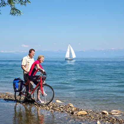 Zwei Radfahrer mit rotem Fahrrad stehen am steinigen Ufer des Bodensees. Weißes Segelboot auf blauem Wasser mit Bergen im Hintergrund.