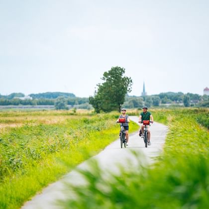 Two cyclists riding on a paved path through green East Frisian countryside with fields, trees, and a church spire visible in the distance.