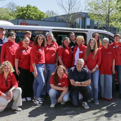 Großes Team-Gruppenfoto mit Personen in roten Shirts, die vor einem weißen Transporter stehen und knien, draußen an einem sonnigen Tag.