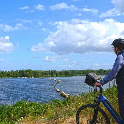 Radfahrer mit Helm steht neben blauem Fahrrad auf Grasweg mit Blick auf IJsselmeer mit Schilf und fernen Inseln unter bewölktem Himmel.