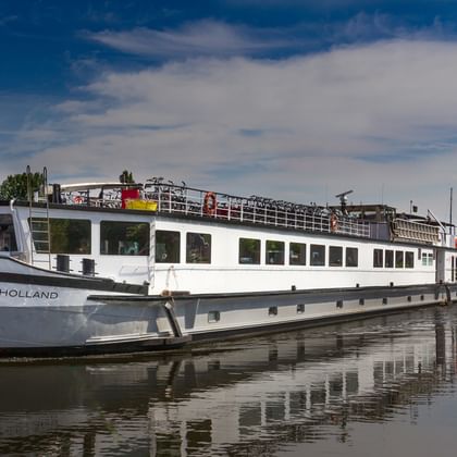White river cruise ship named De Holland moored on calm water with reeds in foreground and blue sky with white clouds above.