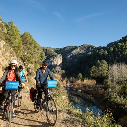 Gruppe von Radfahrern auf einem Flussweg in der Region Valencia, mit Felsklippen und grünen Hügeln unter blauem Himmel.