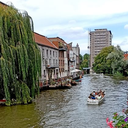 Touristenboot auf Kanal in Brügge mit historischen Gebäuden, Trauerweide und modernem Turm im Hintergrund. Bunte Blumen im Vordergrund sichtbar.