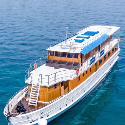 Luftaufnahme der MY Linda, ein weißes Kreuzfahrtschiff mit Holzdeck und blauen Sonnenliegen auf dem Dach, vor Anker in türkisfarbenem Wasser.