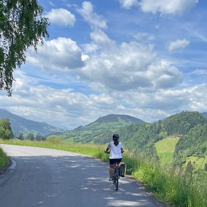 Radfahrer auf asphaltierter Straße entlang der Alpe Adria Route mit grünen Hügeln, Wäldern und Bergen unter blauem Himmel mit weißen Wolken.