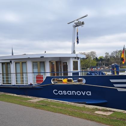 Blau-weißes Flusskreuzfahrtschiff MS Casanova am grasbewachsenen Flussufer unter bewölktem Himmel, mit Bäumen und Gebäuden im Hintergrund.