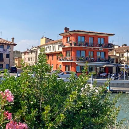 Uferblick auf Marano in Friaul mit bunten Gebäuden, Kirchturm, Kanal mit Booten und rosa blühenden Büschen im Vordergrund.