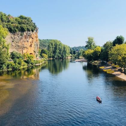 Blick auf die Dordogne mit Kanus auf dem Wasser, Sandstränden an beiden Ufern, üppigen grünen Bäumen und einer Kalksteinklippe links.