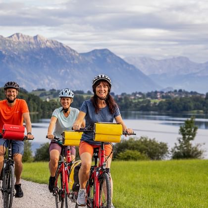 Drei Radfahrer mit Helmen fahren auf einem Weg entlang eines Sees in Österreich. Berge und grüne Wiesen im Hintergrund unter bewölktem Himmel.