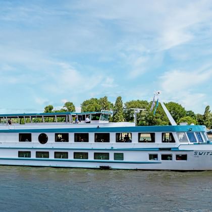 Weißes Flusskreuzfahrtschiff MS Switzerland mit blauem Zierstreifen auf ruhigem Wasser. Bäume und blauer Himmel im Hintergrund.