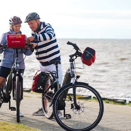 Zwei Radfahrer mit Helmen schauen gemeinsam auf ein Handy an einem Küstenweg. Ihre Tourenräder mit Packtaschen stehen daneben, Meer im Hintergrund.