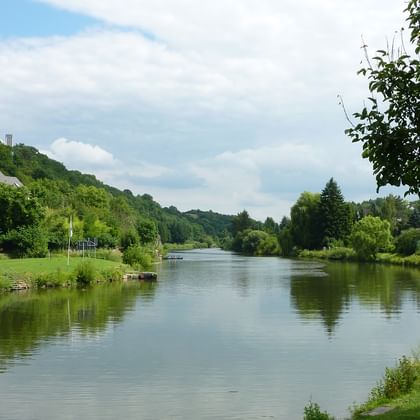 Ruhige Lahn mit grünen Wiesen und Bäumen an beiden Ufern. Häuser in der Ferne unter bewölktem Himmel mit blauen Stellen.