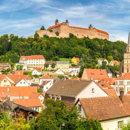 Panoramablick auf Kulmbach mit historischer Burg auf dem Hügel über der Stadt mit roten Ziegeldächern, Kirchturm und grünen Wäldern.
