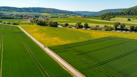 Luftaufnahme des Weserberglandes mit asphaltiertem Radweg durch grüne Felder, gelbes Rapsfeld und Fluss mit Bäumen.